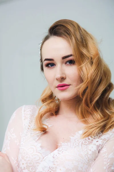 Young attractive bride with blond hair with wedding makeup and hairstyle in a white lace peignoir in a bright interior