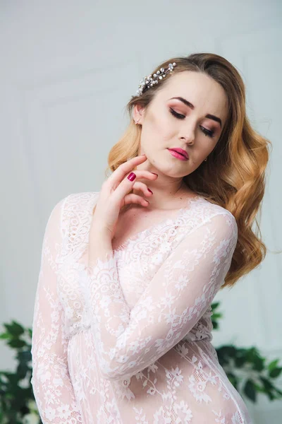 Young attractive bride with blond hair with wedding makeup and hairstyle in a white lace peignoir in a bright interior