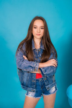 A cute teenage girl with long hair in trendy denim youth clothes and a red T-shirt on a blue background in the studio. Teenage fashion