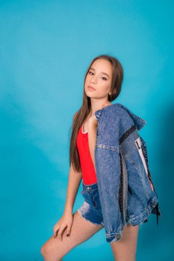 A cute teenage girl with long hair in trendy denim youth clothes and a red T-shirt on a blue background in the studio. Teenage fashion