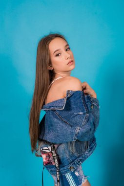 A cute teenage girl with long hair in trendy denim youth clothes and a red T-shirt on a blue background in the studio. Teenage fashion