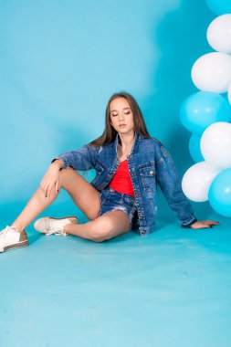 A cute teenage girl with long hair in trendy denim youth clothes and a red T-shirt on a blue background with a blueand white ballons in the studio. Teenage fashion
