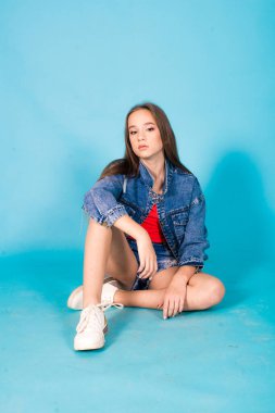 A cute teenage girl with long hair in trendy denim youth clothes and a red T-shirt on a blue background in the studio. Teenage fashion