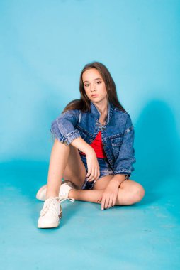 A cute teenage girl with long hair in trendy denim youth clothes and a red T-shirt on a blue background in the studio. Teenage fashion