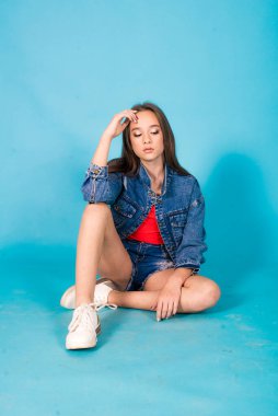 A cute teenage girl with long hair in trendy denim youth clothes and a red T-shirt on a blue background in the studio. Teenage fashion