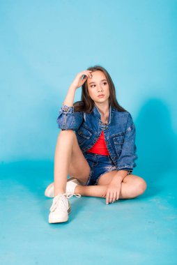 A cute teenage girl with long hair in trendy denim youth clothes and a red T-shirt on a blue background in the studio. Teenage fashion