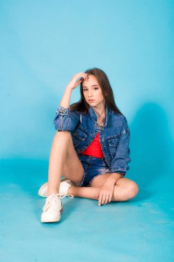 A cute teenage girl with long hair in trendy denim youth clothes and a red T-shirt on a blue background in the studio. Teenage fashion