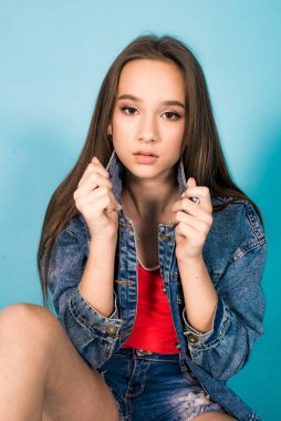 A cute teenage girl with long hair in trendy denim youth clothes and a red T-shirt on a blue background in the studio. Teenage fashion