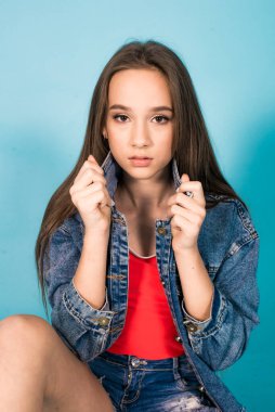 A cute teenage girl with long hair in trendy denim youth clothes and a red T-shirt on a blue background in the studio. Teenage fashion