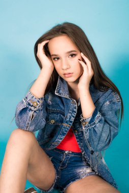 A cute teenage girl with long hair in trendy denim youth clothes and a red T-shirt on a blue background in the studio. Teenage fashion
