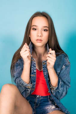 A cute teenage girl with long hair in trendy denim youth clothes and a red T-shirt on a blue background in the studio. Teenage fashion