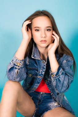 A cute teenage girl with long hair in trendy denim youth clothes and a red T-shirt on a blue background in the studio. Teenage fashion