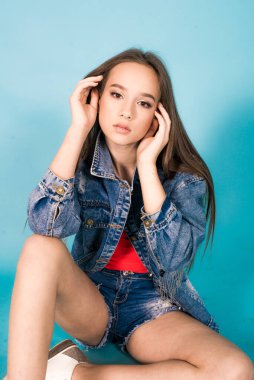 A cute teenage girl with long hair in trendy denim youth clothes and a red T-shirt on a blue background in the studio. Teenage fashion