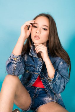 A cute teenage girl with long hair in trendy denim youth clothes and a red T-shirt on a blue background in the studio. Teenage fashion