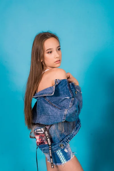 A cute teenage girl with long hair in trendy denim youth clothes and a red T-shirt on a blue background in the studio. Teenage fashion