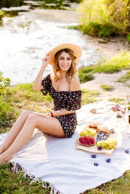 Young attractive girl with dark hair in a summer dress on a picnic in nature by the river in summer at sunset. Healthy life.