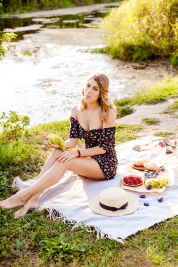 Young attractive girl with dark hair in a summer dress on a picnic in nature by the river in summer at sunset. Healthy life.