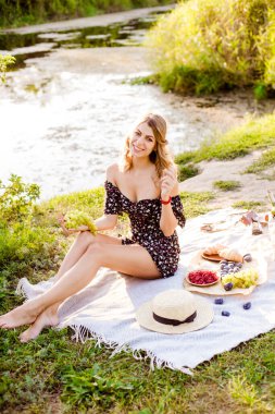 Young attractive girl with dark hair in a summer dress on a picnic in nature by the river in summer at sunset. Healthy life.