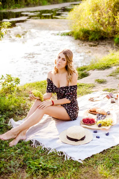 Young attractive girl with dark hair in a summer dress on a picnic in nature by the river in summer at sunset. Healthy life.