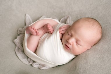 A cute baby sleeps wrapped in an airy milk-colored blanket on a beige plaid on a bed at home.