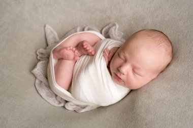 A cute baby sleeps wrapped in an airy milk-colored blanket on a beige plaid on a bed at home.