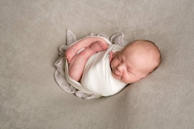 A cute baby sleeps wrapped in an airy milk-colored blanket on a beige plaid on a bed at home.
