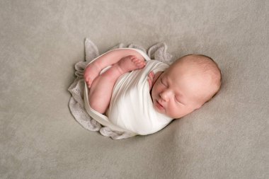 A cute baby sleeps wrapped in an airy milk-colored blanket on a beige plaid on a bed at home.