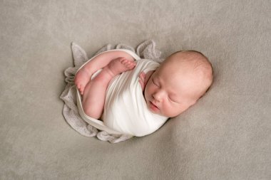 A cute baby sleeps wrapped in an airy milk-colored blanket on a beige plaid on a bed at home.