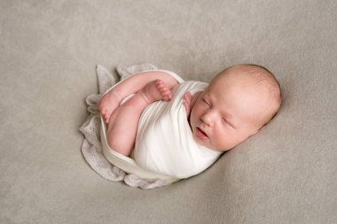 A cute baby sleeps wrapped in an airy milk-colored blanket on a beige plaid on a bed at home.