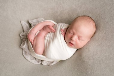 A cute baby sleeps wrapped in an airy milk-colored blanket on a beige plaid on a bed at home.