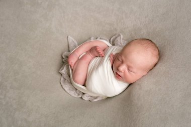 A cute baby sleeps wrapped in an airy milk-colored blanket on a beige plaid on a bed at home.