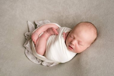 A cute baby sleeps wrapped in an airy milk-colored blanket on a beige plaid on a bed at home.