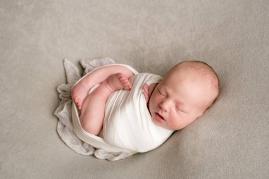 A cute baby sleeps wrapped in an airy milk-colored blanket on a beige plaid on a bed at home.