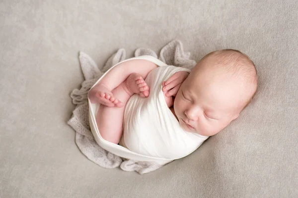 A cute baby sleeps wrapped in an airy milk-colored blanket on a beige plaid on a bed at home.