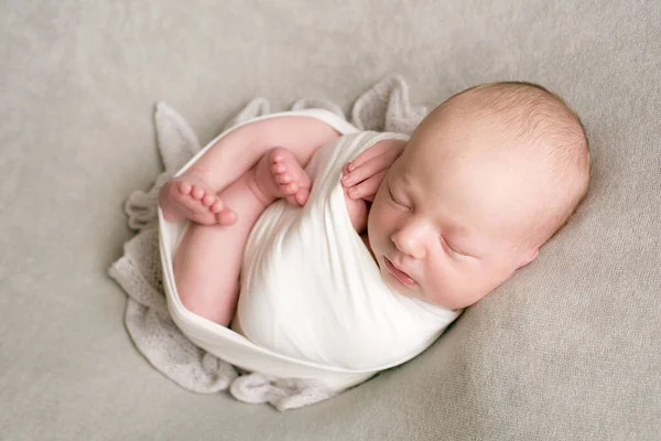 A cute baby sleeps wrapped in an airy milk-colored blanket on a beige plaid on a bed at home.