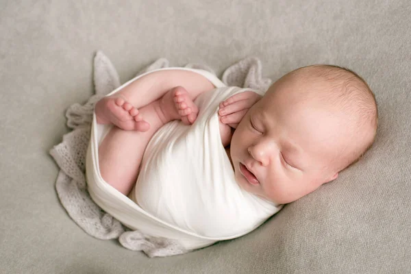A cute baby sleeps wrapped in an airy milk-colored blanket on a beige plaid on a bed at home.