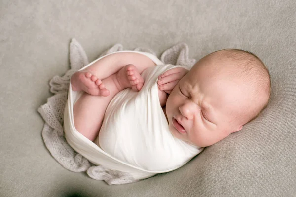 A cute baby sleeps wrapped in an airy milk-colored blanket on a beige plaid on a bed at home.