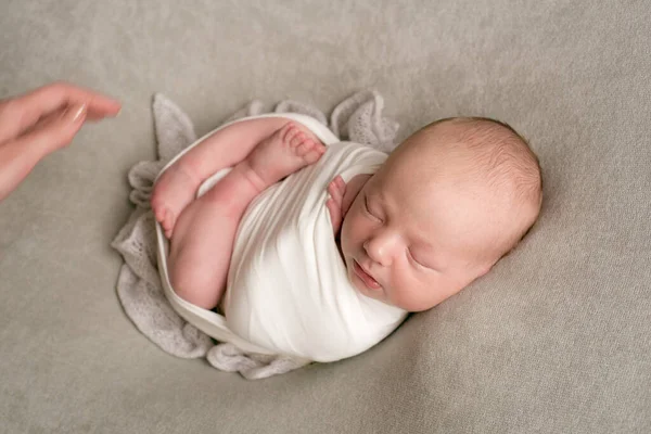 A cute baby sleeps wrapped in an airy milk-colored blanket on a beige plaid on a bed at home.
