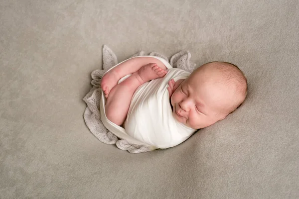 A cute baby sleeps wrapped in an airy milk-colored blanket on a beige plaid on a bed at home.
