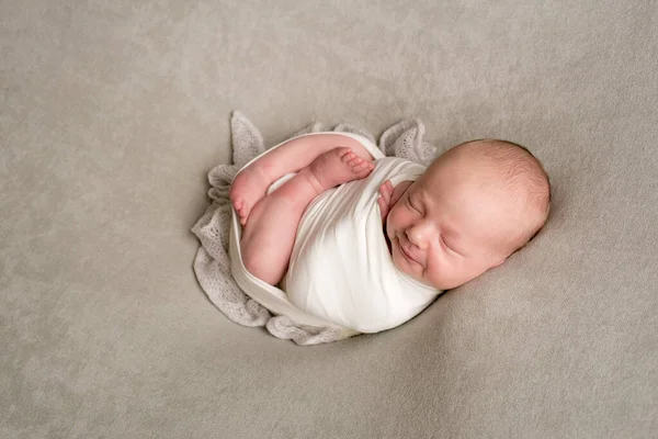 A cute baby sleeps wrapped in an airy milk-colored blanket on a beige plaid on a bed at home.