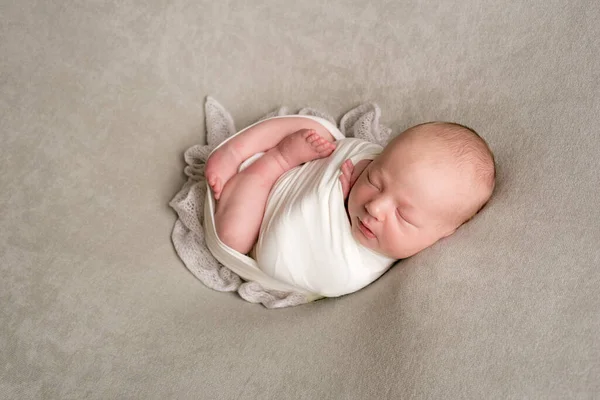 A cute baby sleeps wrapped in an airy milk-colored blanket on a beige plaid on a bed at home.
