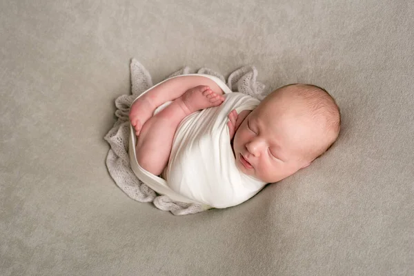 A cute baby sleeps wrapped in an airy milk-colored blanket on a beige plaid on a bed at home.
