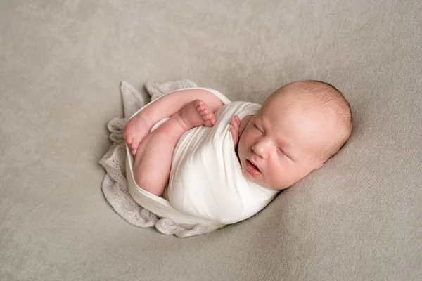 A cute baby sleeps wrapped in an airy milk-colored blanket on a beige plaid on a bed at home.
