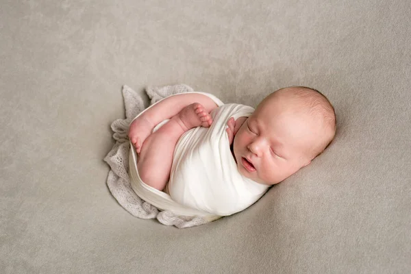 A cute baby sleeps wrapped in an airy milk-colored blanket on a beige plaid on a bed at home.
