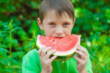 A cute boy in a green t-shirt eats a juicy ripe watermelon in the summer in the garden. Healthy lifestyle. Happy childhood