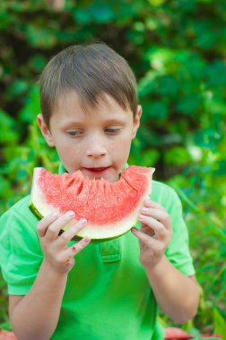 A cute boy in a green t-shirt eats a juicy ripe watermelon in the summer in the garden. Healthy lifestyle. Happy childhood
