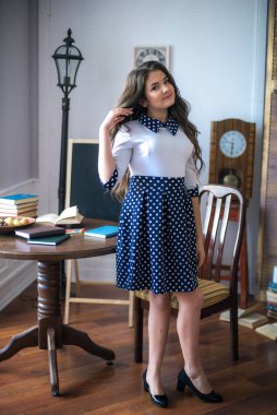 A cute teenage girl with long hair in a classic school dress in a classic interior with books and notebooks at the lesson. School life. Back to school