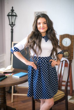 A cute teenage girl with long hair in a classic school dress in a classic interior with books and notebooks at the lesson. School life. Back to school