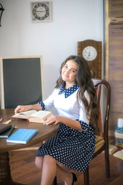 A cute teenage girl with long hair in a classic school dress in a classic interior with books and notebooks at the lesson. School life. Back to school
