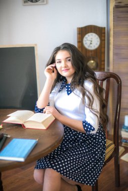 A cute teenage girl with long hair in a classic school dress in a classic interior with books and notebooks at the lesson. School life. Back to school